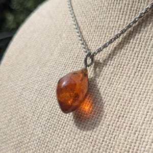 May include: A silver chain necklace with a teardrop-shaped amber pendant. The amber pendant is a warm, translucent orange-brown colour. The chain has a woven design. The pendant is suspended from a small silver loop.