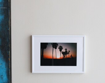 Puede incluir: Una fotografía enmarcada de una hilera de palmeras silueteada contra un cielo de puesta de sol. La foto está colgada en una pared blanca con una escultura de metal oxidado y una planta en maceta en primer plano.