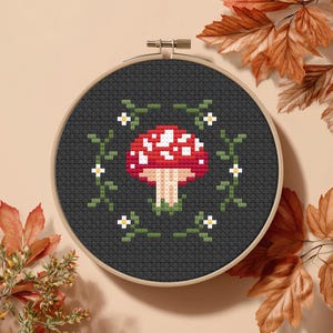 May include: A cross-stitch embroidery hoop with a red and white mushroom design. The mushroom is encircled by a green vine with small white flowers, set against a dark grey background. The hoop is displayed with autumn leaves.
