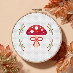 May include: A cross-stitch embroidery hoop featuring a red mushroom with white spots and a bow. Green sprigs with red berries surround the mushroom. The embroidery is set against a white background within a wooden hoop, with autumn leaves in the background.