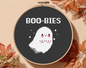 May include: A cross-stitch hoop featuring a pixelated white ghost with pink cheeks and the word "BOO-BIES" in white block letters on a black background. The hoop is set against a neutral background with autumn leaves.