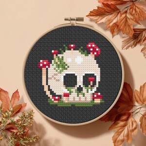 May include: Cross-stitch embroidery of a skull decorated with red and white mushrooms and green foliage. The skull is off-white with black eye sockets and nose. The embroidery is set in a black hoop, with a beige background and autumn leaves.