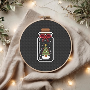 Op de afbeelding: Een borduurring met kruissteek met een zwarte achtergrond en een potontwerp. In de pot zit een kerstboom met een rode strik, een kurken dop en een witte basis. De borduurring is van hout.