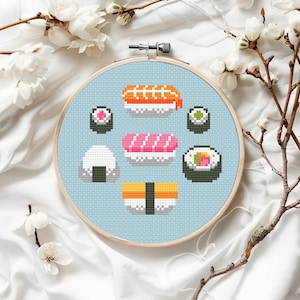 Puede incluir: Un bastidor de bordado de punto de cruz con varias piezas de sushi en estilo pixelado. El sushi incluye nigiri, rollos maki y onigiri, todo en colores vibrantes sobre un fondo azul claro. El bastidor de madera está rodeado de flores blancas.