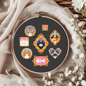 May include: A cross-stitch embroidery hoop featuring a dark gray fabric with several colorful pixelated cat-themed designs. The designs include cat faces, a fishbone, and a heart. The hoop is set against a neutral fabric background.