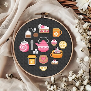 Op de afbeelding: Een borduurring met kruissteek met een zwarte achtergrond, met kleurrijke thee- en desserticonen. Het ontwerp omvat een roze theepot, stukken cake en theekopjes. De ring is geplaatst tegen een neutrale stof en een bloemstuk.