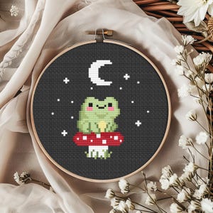 Peut inclure: Broderie au point de croix dans un cercle en bois. Le motif représente une grenouille verte assise sur un champignon rouge à pois blancs, sous un croissant de lune blanc et des étoiles sur fond noir. La broderie est entourée d'un tissu neutre et de fleurs blanches.