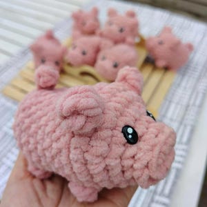 Può includere: Una collezione di giocattoli a forma di maiale rosa fatti a mano all'uncinetto. I giocattoli a forma di maiale hanno piccoli occhi neri e sono realizzati con un filato morbido e strutturato. Diversi maiali sono disposti su una superficie di legno, con un maiale in primo piano.