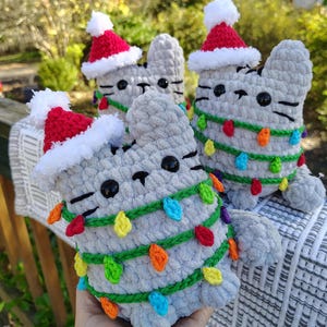 May include: Crocheted gray cat plushies wearing red Santa hats and wrapped in colorful Christmas lights. The cat toys have black eyes and whiskers. The plushies are handmade and suitable for holiday decor.