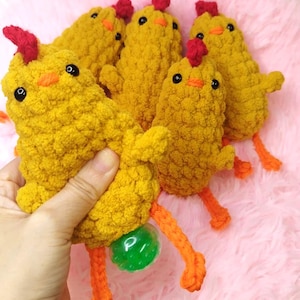Puede incluir: Una colección de juguetes de crochet amarillos hechos a mano con crestas rojas, picos naranjas y patas naranjas. Los pollos tienen ojos negros y se exhiben sobre una superficie rosa. Un pollo tiene una bola verde unida a sus patas.