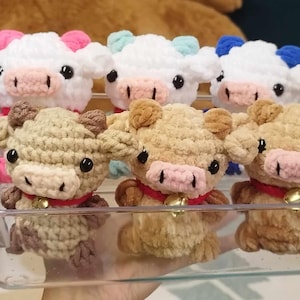 May include: A selection of handmade crochet cow plushies in a clear plastic container. The cows are white, tan, and brown with pink, blue, and brown accents. Each cow has a red collar and a small gold bell.