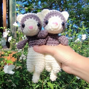 Puede incluir: Dos peluches de hurón hechos a mano con ganchillo. Los hurones tienen cuerpos blancos, marcas grises-moradas y narices rosas. Se sostienen frente a un fondo de follaje verde y flores blancas.
