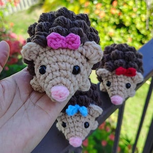 May include: Three crocheted hedgehog plushies with different colored bows. The largest hedgehog has a pink bow, the middle hedgehog has a blue bow, and the smallest hedgehog has a red bow.