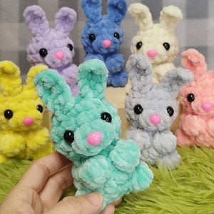 May include: A collection of handmade plush bunnies in various pastel colors, including yellow, lavender, blue, and pink. Each bunny has black eyes and a pink nose. The bunnies are made of a soft, textured material.