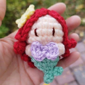 May include: A handmade crochet doll of a mermaid with red hair, a yellow flower, and a purple bow. The doll has a light skin tone and a green tail. The doll is made of yarn and is a cute, whimsical design.