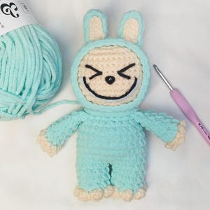 May include: A handmade crochet bunny doll in a light blue jumpsuit with a cream-colored face and accents. The bunny has a smiling face with black embroidered details. A ball of light blue yarn and a crochet hook are also in the image.