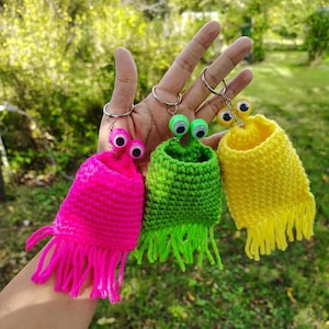 Può includere: Tre portachiavi all'uncinetto fatti a mano in colori vivaci: rosa, verde e giallo. Ogni portachiavi presenta un corpo cilindrico con frange e occhi mobili. I portachiavi sono tenuti su uno sfondo verde sfocato.