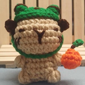 May include: A handmade crochet animal figure in beige, green, and brown. The figure has a green hat, brown ears, and is holding an orange fruit with a green stem. The figure is made of yarn and has a whimsical design.