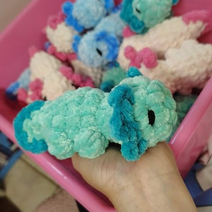 May include: A close-up of a turquoise and blue crochet axolotl plush toy. The plush toy has a textured, fuzzy appearance with blue gills and a small black eye. Other similar plush toys are in a pink bin.