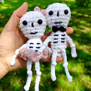 Puede incluir: Dos muñecos de esqueleto de ganchillo hechos a mano. Uno tiene un lazo rosa y un tutú rosa, el otro una pajarita negra. Ambos son blancos con ojos y sonrisas negras. Los muñecos se sostienen sobre un fondo verde.