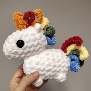 May include: A handmade white crochet unicorn with a black eye and a colorful mane and tail. The mane features orange, red, and yellow accents, while the tail showcases a rainbow of colors. This plush toy is a whimsical and cuddly creation.