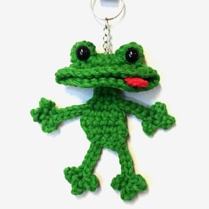 May include: A handmade green crochet frog keychain with black button eyes and a red tongue. The frog has outstretched arms and legs, and is attached to a silver keyring. The frog is made of a textured yarn.