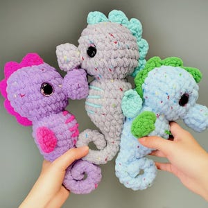 May include: Three handmade crochet seahorse plush toys in shades of purple, gray, and blue. Each plush has a textured body with colorful flecks, a contrasting fin, and large, shiny black eyes. The seahorses are held up against a gray background.