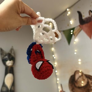 May include: A small, handmade crochet ornament featuring a red and blue spherical character head with white felt eyes, attached to a white crocheted web-like structure. The item is crafted from yarn and designed as a hanging decorative piece.