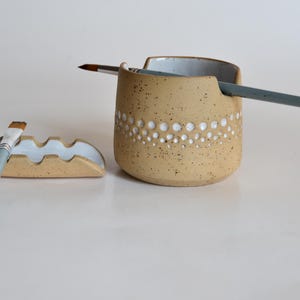 Pode incluir: Um porta-pincéis de cerâmica bege salpicado com uma fileira de pontos brancos e um pequeno descanso de pincel a condizer. Dois pincéis são colocados no suporte. O suporte de material de arte é um item funcional e decorativo.