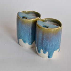 Può includere: Due tazze in ceramica dal design unico. Le tazze sono cilindriche con una glassa blu che cola su una base bianca. Il bordo superiore è di un colore oro a contrasto. Ogni tazza ha un'apertura curva sul lato.
