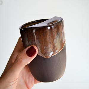 Könnte beinhalten: Eine handgefertigte Keramiktasse mit einem einzigartigen Design. Die obere Hälfte der Tasse ist glänzend braun mit Streifen aus hellem Braun und Beige, während die untere Hälfte matt dunkelbraun ist. Die Tasse hat einen markanten kantigen Rand.