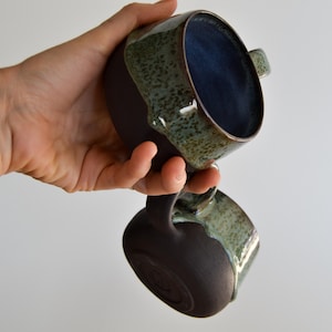 Könnte beinhalten: Zwei Keramikbecher mit Henkeln. Die Becher sind dunkelbraun mit einer gesprenkelten grün-blauen Glasur am Rand und einem tiefblauen Innenraum. Die Becher werden in einer Hand vor weißem Hintergrund gehalten.