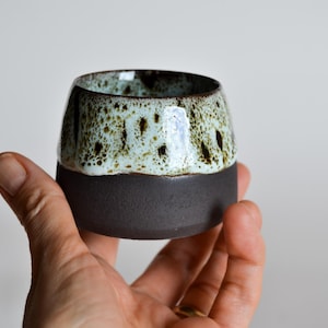 Può includere: Una piccola tazza in ceramica fatta a mano con una base marrone scuro e una parte superiore smaltata maculata verde chiaro e marrone. La tazza è tenuta in mano, mostrando il suo design unico e organico. Ideale per caffè o tè.
