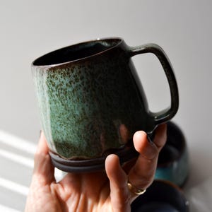 Könnte beinhalten: Eine handgefertigte Keramiktasse mit dunkelgrüner Glasur und braunem Rand. Die Tasse hat einen großen Henkel und ein leicht abgewinkeltes Design. Die Tasse wird in einer Hand gehalten, eine weitere Tasse ist im Hintergrund zu sehen.