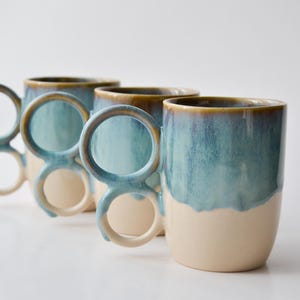 May include: Three ceramic mugs with unique handles. The mugs have a two-tone design, with a cream-colored base and a blue-green glaze on the upper half. The handles are a distinctive design, with two circular openings. The mugs are arranged in a diagonal line.