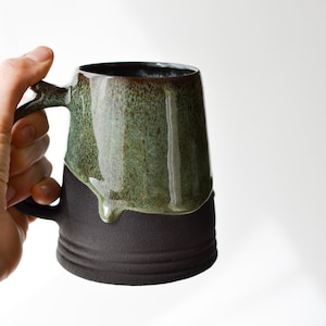 May include: A large, ceramic mug with a black and green glaze. The glaze has a drip effect, with the green glaze dripping down the side of the mug.