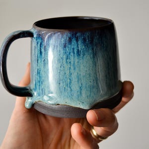 May include: A blue and black ceramic mug with a drip glaze. The mug has a rounded body and a handle.