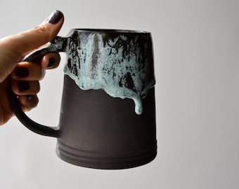 Könnte beinhalten: Ein schwarzer Bierkrug aus Keramik mit einem hellblauen Überzug, der an der Seite heruntertropft. Der Krug hat einen Henkel und einen breiten Boden.