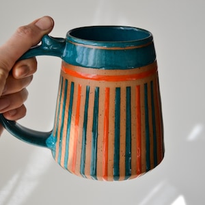 Può includere: Una tazza in ceramica con manico e bordo verde acqua e corpo beige. Il corpo è decorato con strisce verticali verde acqua e arancioni. La tazza è tenuta da una mano.