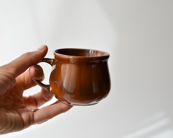 May include: A brown ceramic mug with a rounded shape and a handle. The mug is held in a hand.