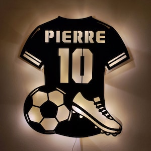 Könnte beinhalten: Ein schwarzes und weißes Holz-Nachtlicht in Form eines Fußballtrikots mit dem Namen "PIERRE" und der Nummer "10" auf der Vorderseite. Ein Fußball und ein Fußballschuh befinden sich unterhalb des Trikots. Das Nachtlicht ist beleuchtet und hat eine Fernbedienung.