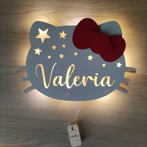 Può includere: Una luce notturna in legno bianco a forma di personaggio Hello Kitty con un fiocco rosso, il nome Valeria ritagliato al centro e stelle intorno ai bordi. La luce è illuminata e ha un piccolo interruttore in legno su un cavo.
