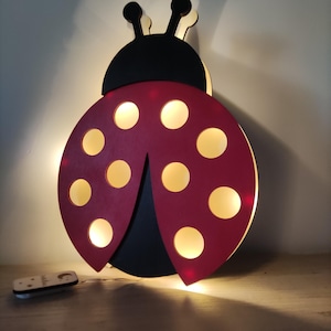 May include: A wooden ladybug-shaped wall light with a red body and black head. The ladybug has yellow circular lights and a remote control. The light is designed for a child's room or nursery.