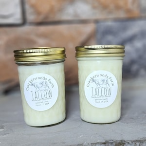 Op de afbeelding: Twee glazen potten met gouden deksels gevuld met witte was. De potten hebben etiketten met de tekst "Crackerwoods Farm Tallow Rendered from natural pastured cattle March 17, 2024" en bevatten een zwart-witte koe-illustratie.