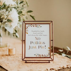 May include: A copper-colored framed sign with a white background and a floral design. The sign reads "Thankful for family, food, and fun! Let's keep the table happy... No Politics Just Pie!"