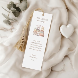 Puede incluir: Un marcapáginas blanco con un borla dorada. El marcapáginas presenta una ilustración de acuarela de tres ositos de peluche sentados en una pila de libros. El texto en el marcapáginas dice "A New Chapter will soon begin."  "Thank you for celebrating with us! Your love and support mean so much as we welcome our little one."  "Anne's Baby Shower 9.17.25"