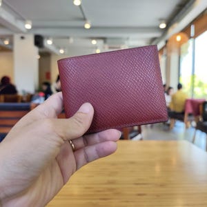 May include: A burgundy leather wallet with a textured surface. The wallet is rectangular and appears to be bi-fold. The image is taken indoors, with a blurred background.