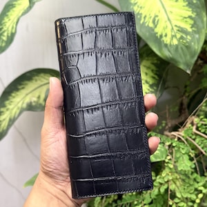 Puede incluir: Una cartera larga y rectangular de color negro con una textura en relieve que imita la piel de cocodrilo. La cartera presenta un acabado liso y brillante, con un perfil estructurado y delgado de bordes rectos y definidos.