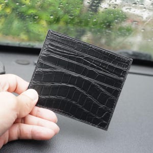 Puede incluir: Un tarjetero negro con un patrón texturizado de piel de cocodrilo. El accesorio rectangular tiene múltiples ranuras horizontales para organizar tarjetas y está construido con un perfil delgado y bordes cosidos a lo largo del perímetro.
