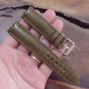 Könnte beinhalten: Ein olivgrünes Uhrenarmband mit einer strukturierten Eidechsenhaut-Optik, tonalen Nähten an den Kanten und einer polierten silberfarbenen Metallschnalle. Das zweiteilige Band verfügt über eine Reihe von Einstelllöchern und zwei Lederschlaufen.